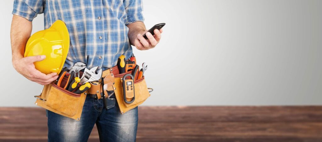 Artisan en chemise à carreaux et jeans, équipé d'une ceinture à outils, consulte son smartphone, un casque jaune à la main.