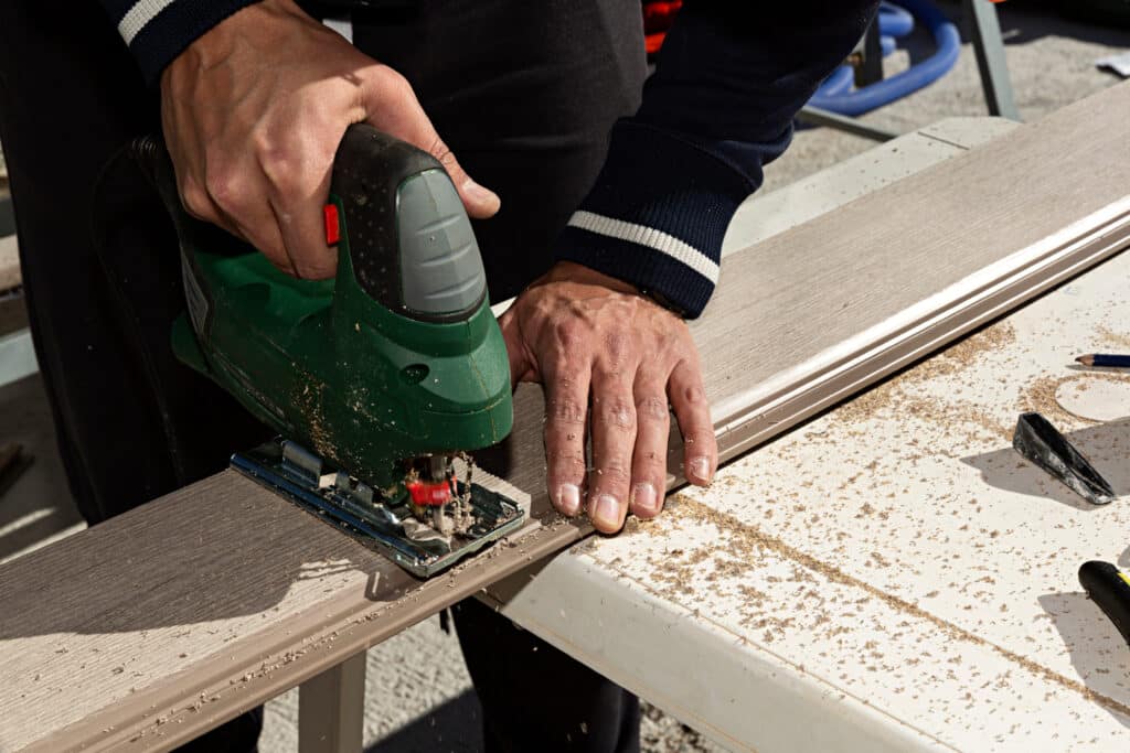 Un homme coupe une planche de terrasse composite avec une scie sauteuse verte, créant de la sciure sur un établi blanc. Travail de précision.