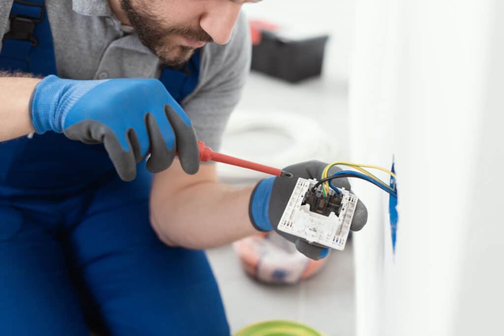 Électricien barbu en gants bleus, concentré sur l'installation d'une prise électrique murale, connectant des fils colorés avec un tournevis.