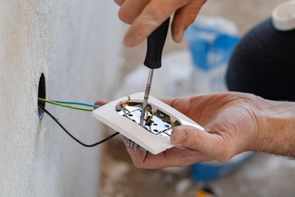 Électricien vissant une prise électrique murale blanche. Câbles (vert/jaune, bleu, noir) sortent d'un trou dans un mur texturé.