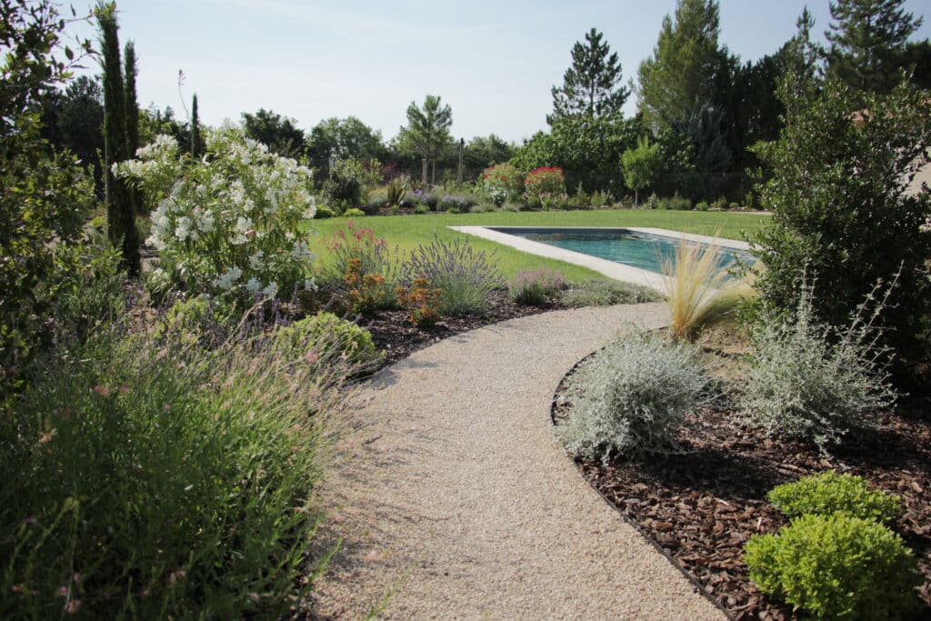 Allée de gravier incurvée menant à une piscine bleue dans un jardin paysager méditerranéen ensoleillé avec lavande et arbustes fleuris.