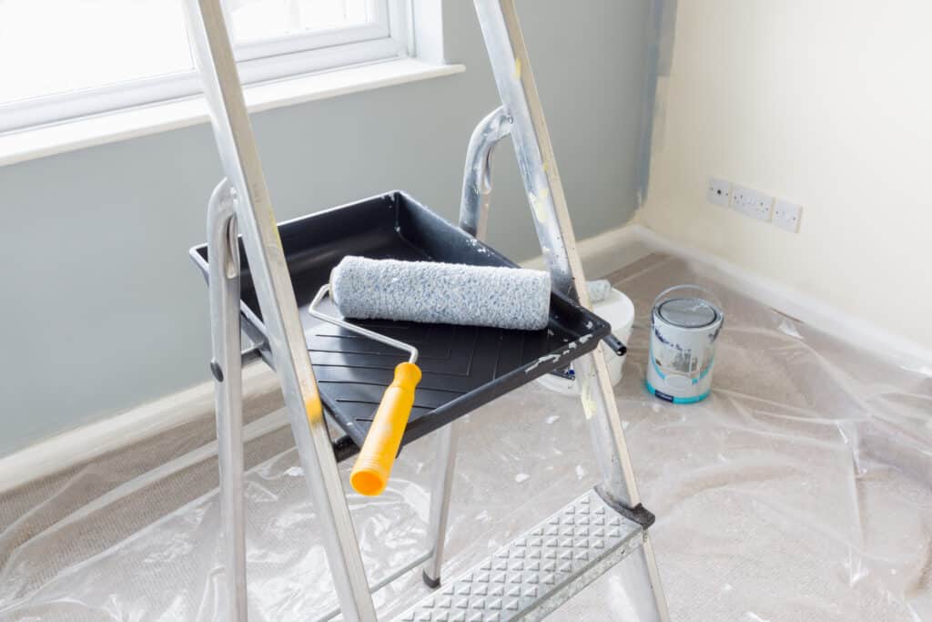 Peinture d'une pièce : échelle, rouleau, bac à peinture sur sol protégé par bâche. Murs en cours de peinture, gris et crème.