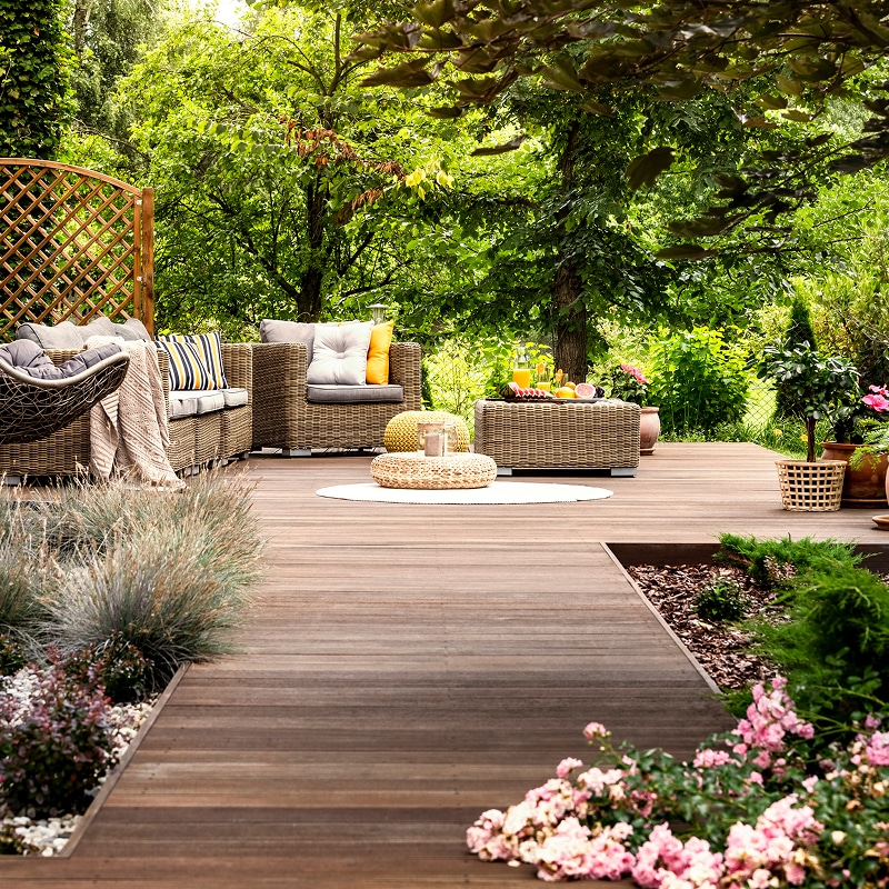 Terrasse en bois avec mobilier en osier dans jardin luxuriant. Coussins colorés, boissons, fruits et fleurs roses.