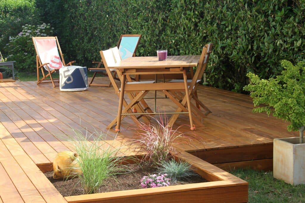 Terrasse en bois clair, salon de jardin et transats (9, 5). Pouf (25), plantes et arbustes. Espace détente ensoleillé.
